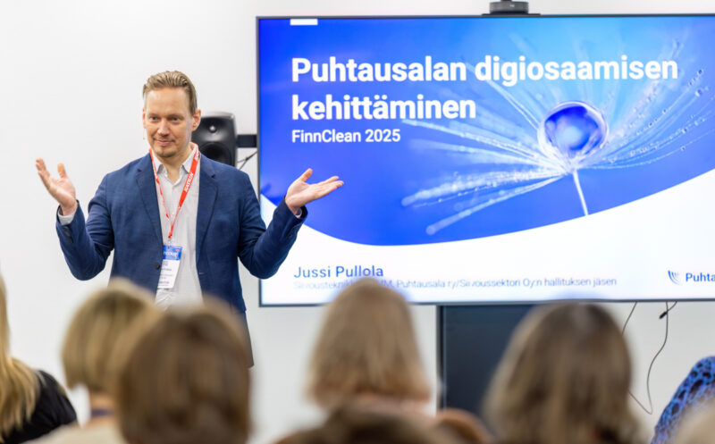 Mies sinisessä pikkutakissa esittelee näytöllä näkyvää esitystä. Hän on kohottanut kätensä näytön suuntaan. Etualalla on yleisöä.
