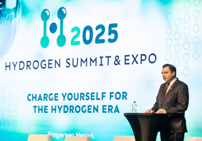 Mies seisoo mustan puhujapöydän takana. Pöydällä on vesipullo. Taustalla on suuri näyttö, jossa on Hydrogen Summit & Expo -tapahtuman logo sekä tekstiä vaaleansinisellä taustalla.